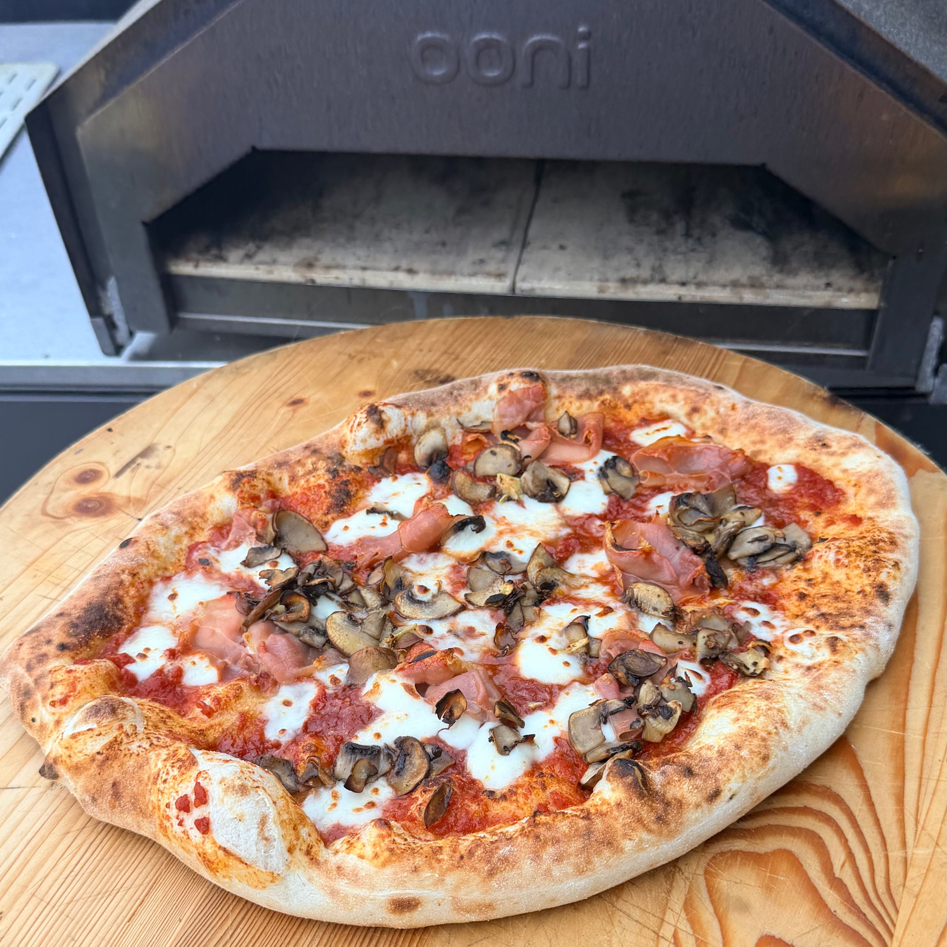 A freshly baked Stir Bakery pizza with mushrooms, ham, and cheese sits on a wooden paddle peel in front of an outdoor oven. 